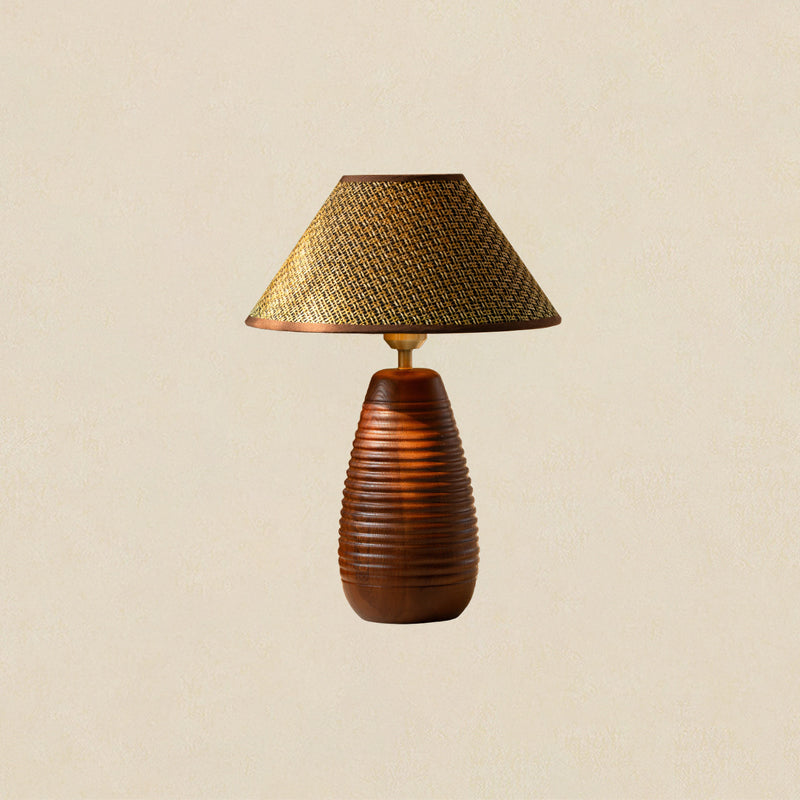 A Clérachic table lamp with a straw weave shade and a tall wood base carved with horizontal ribs, on a beige backdrop.