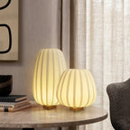 A tall and a round Clérachic table lamp glow side by side on a marble-topped round table in front of linen curtains.