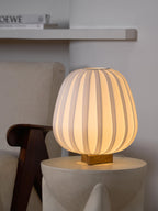 On a cream stone side table beside a wooden-armed chair, this Clérachic table lamp glows softly through its ribbed fabric shade.