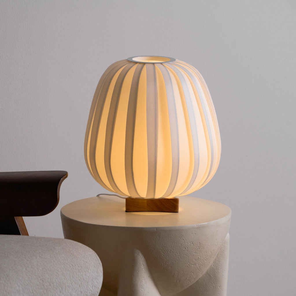 Front view of the round Clérachic table lamp lit warm on a stone side table, its wooden base visible beneath the ribbed shade.