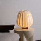 Seen slightly from the side, a lit Clérachic table lamp rests on a stone side table next to a chair with a dark wooden arm.