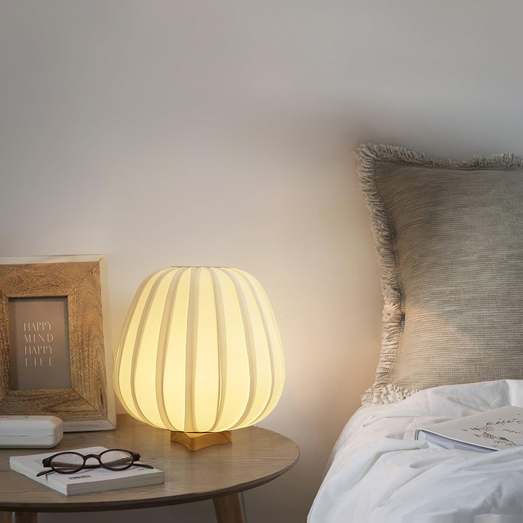 A round Clérachic table lamp glows on a wooden bedside table by a bed with a grey cushion, a framed note and a pair of glasses.