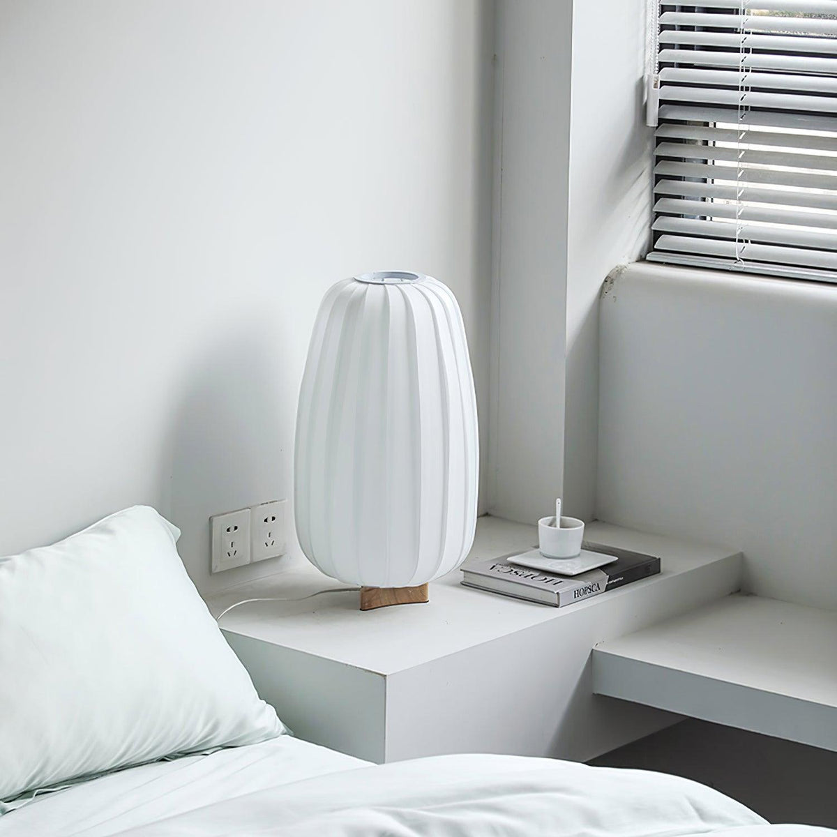 A tall white Clérachic table lamp, unlit, stands on a white bedside shelf beside a book, with a window blind behind it.
