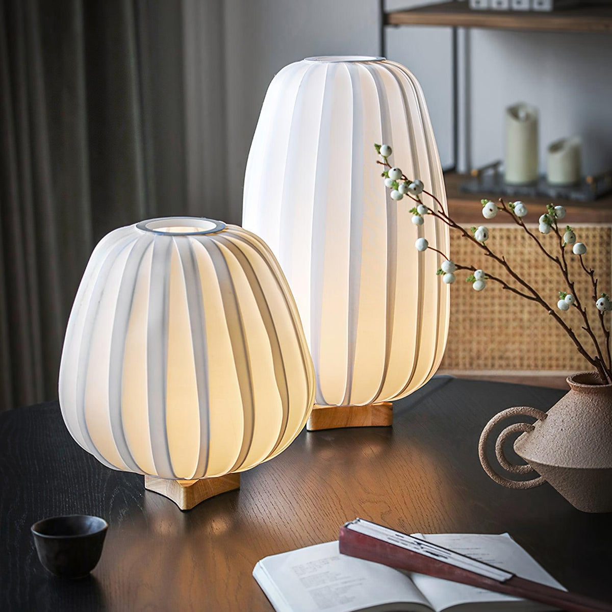 On a dark wood table with a book and a clay vase of white berries, two unlit Clérachic lamps show their ribbed white shades.
