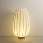 A tall Clérachic fabric table lamp glows on its wooden base against a plain beige wall on a pale wood surface.