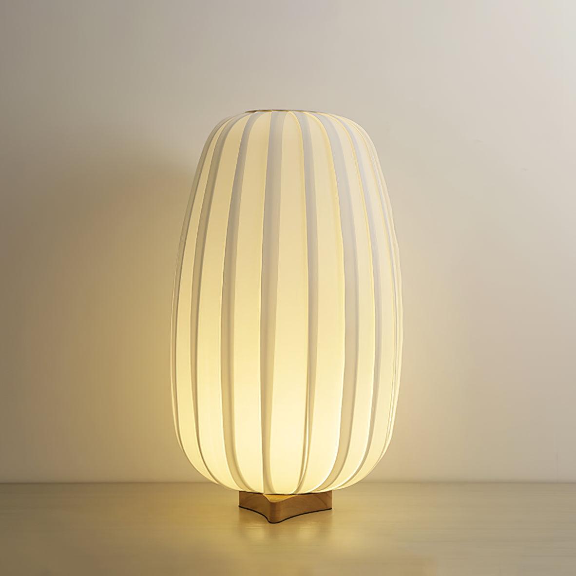 A tall Clérachic fabric table lamp glows on its wooden base against a plain beige wall on a pale wood surface.