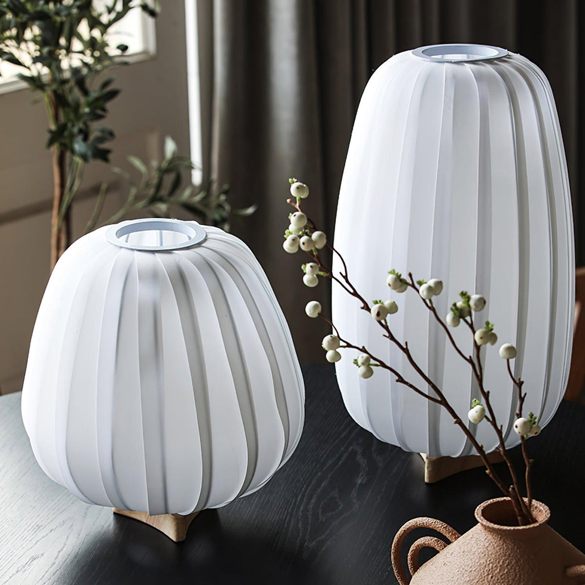 Beside a vase of white berry stems, two unlit Clérachic table lamps show their white ribbed shades on a dark wooden table.