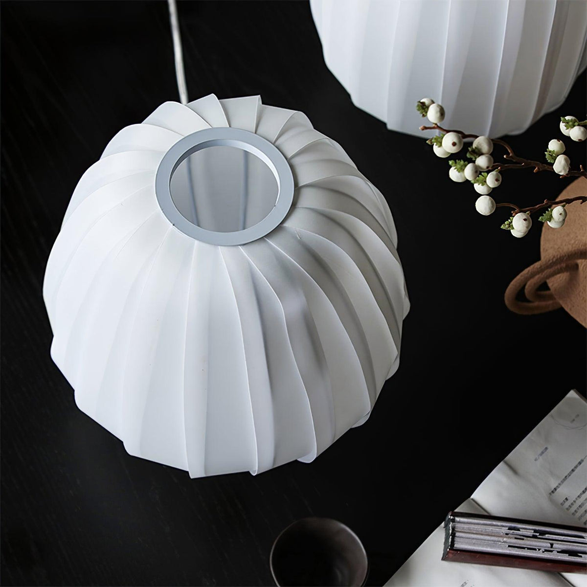 Top-down view of an unlit Clérachic shade, its ribbed white fabric gathering at a round opening, on a black table with white berries.
