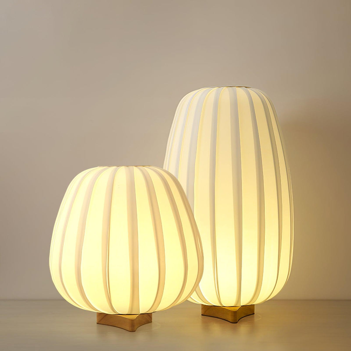 The round and tall Clérachic table lamps glow together on a pale wooden shelf in front of a warm beige wall.