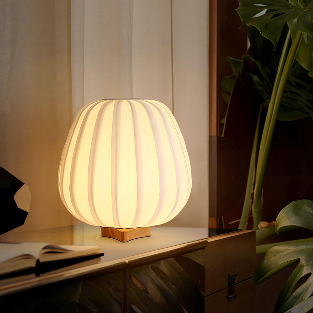 A lit round Clérachic table lamp sits on a low wooden shelf with an open book beside a leafy plant and sheer curtain.