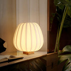 A lit round Clérachic table lamp sits on a low wooden shelf with an open book beside a leafy plant and sheer curtain.