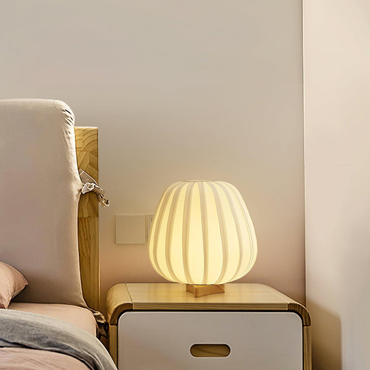 On a white bedside cabinet beside a bed, this round Clérachic table lamp lights the wall behind it with a warm glow.