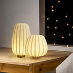Two lit Clérachic table lamps, one tall and one round, stand on a wooden desk by a window with string lights behind.