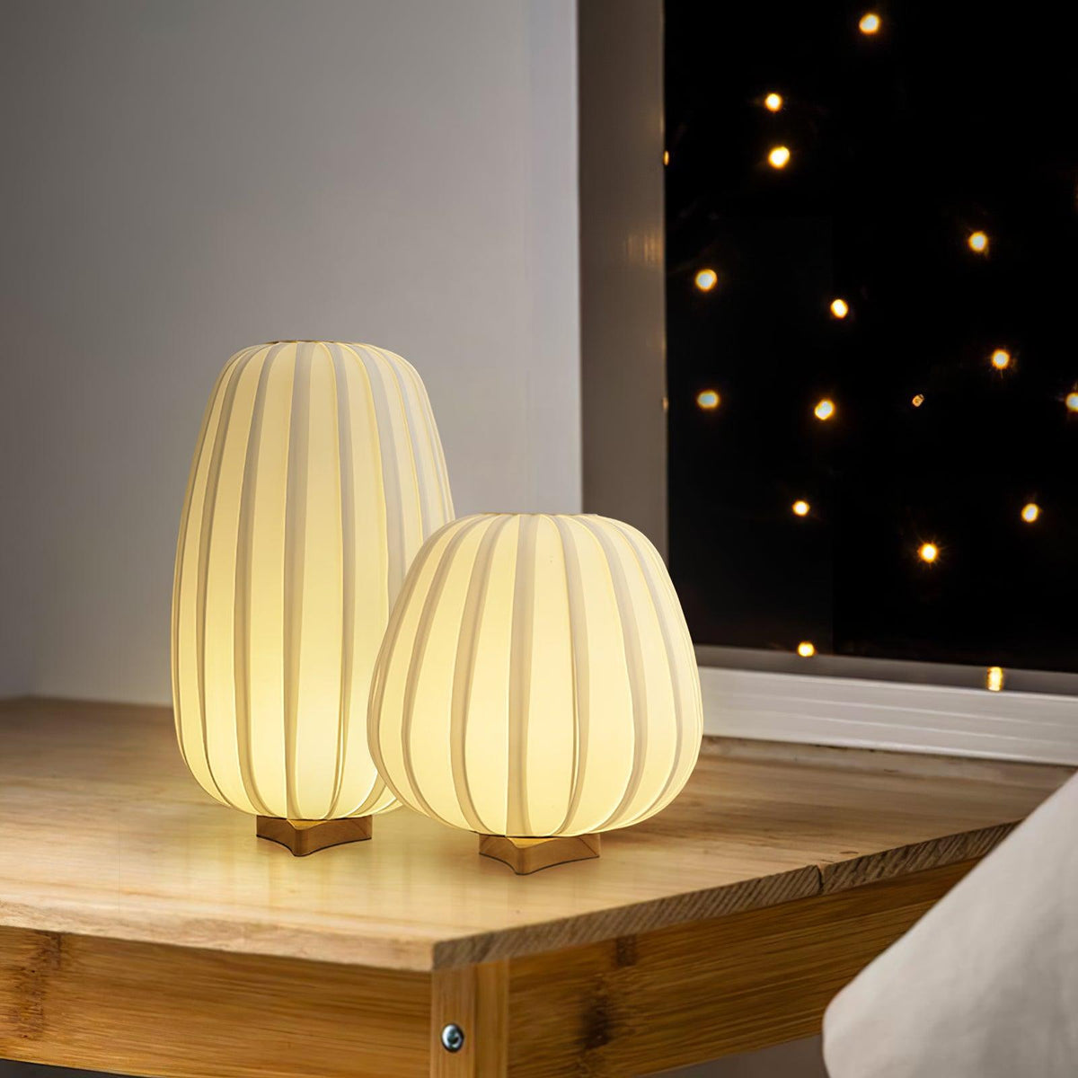 Two lit Clérachic table lamps, one tall and one round, stand on a wooden desk by a window with string lights behind.