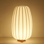 Lit warm gold, this tall Clérachic table lamp on its wooden base is shown against a rich amber backdrop.