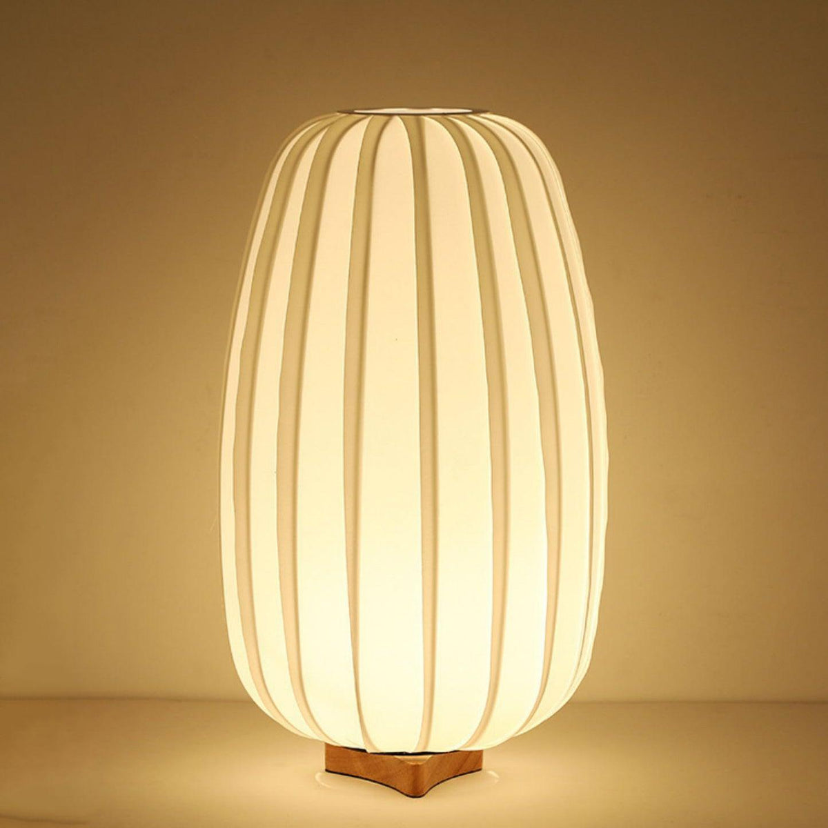 Lit warm gold, this tall Clérachic table lamp on its wooden base is shown against a rich amber backdrop.