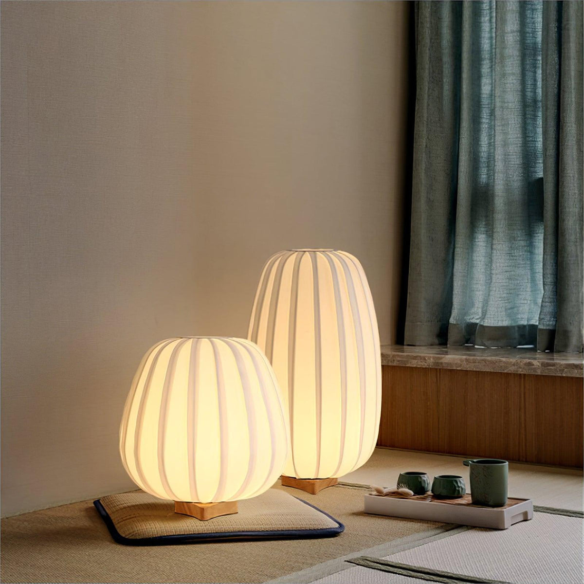 A round and a tall Clérachic lamp glow on a tatami-style mat beside a tea set, with teal curtains at the window.