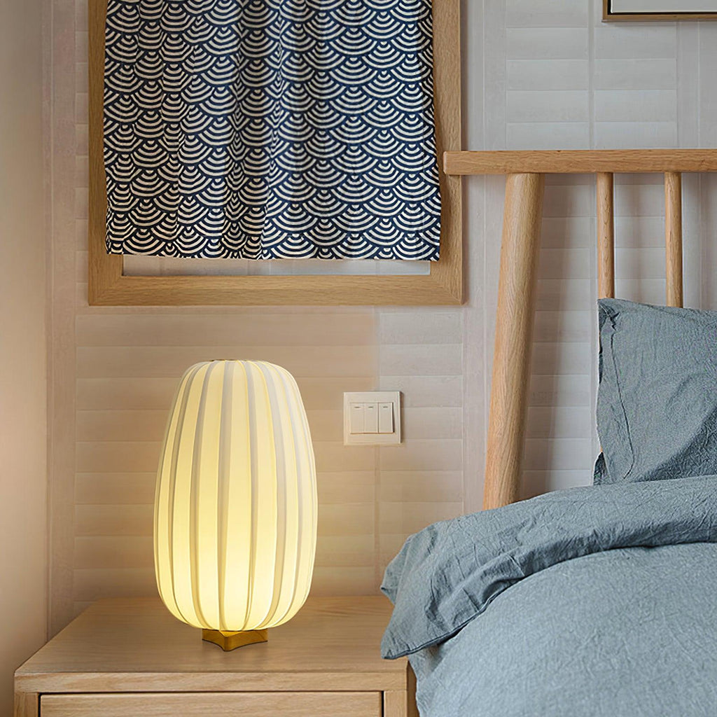 Beside a bed with blue-grey linen and a wood headboard, a tall Clérachic table lamp glows on a pale wood bedside table.