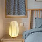 Beside a bed with blue-grey linen and a wood headboard, a tall Clérachic table lamp glows on a pale wood bedside table.