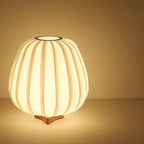 The round Clérachic table lamp glows warm on a wooden base, showing its ribbed fabric shade on an amber backdrop.