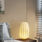 A tall Clérachic table lamp glows on a dark bedside table beside a bed with soft pillows and a slatted window blind.
