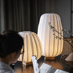 A person reads in the foreground while a round and a tall Clérachic table lamp glow on a dark table beside a vase.