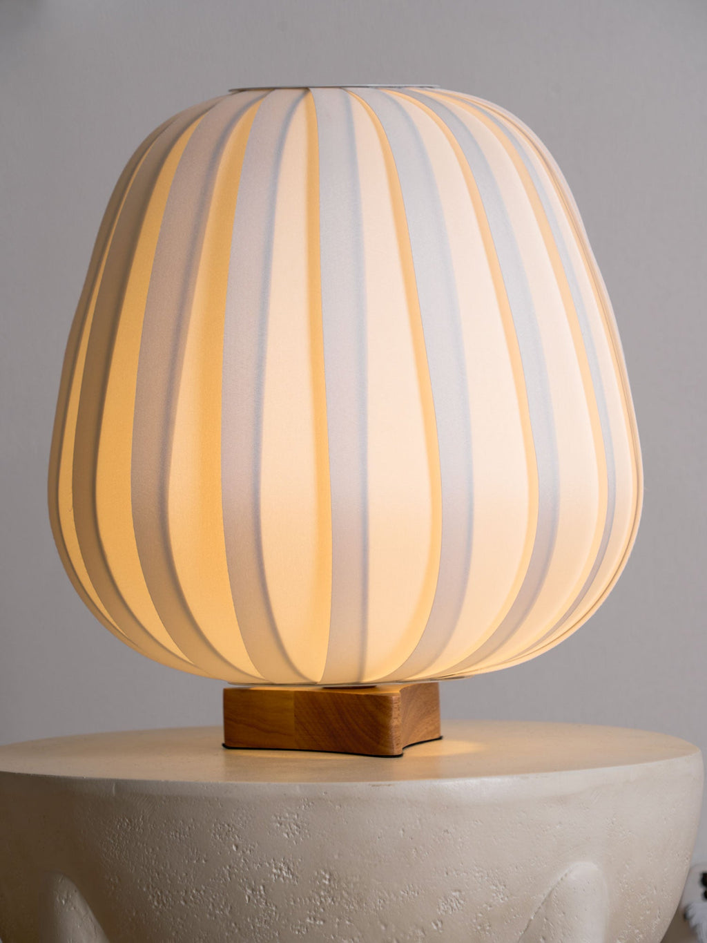 A lit Clérachic table lamp rests on a stone side table, its ribbed fabric shade glowing warm against a grey wall.