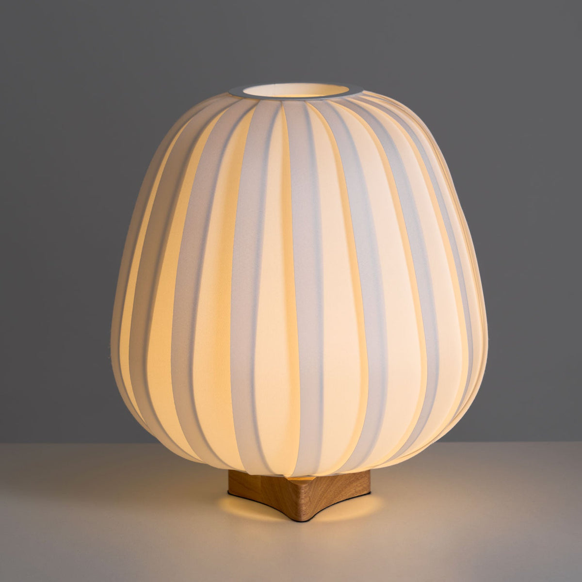 Warm light glows through the ribbed fabric of this Clérachic table lamp, with its top opening and wooden base on a grey surface.