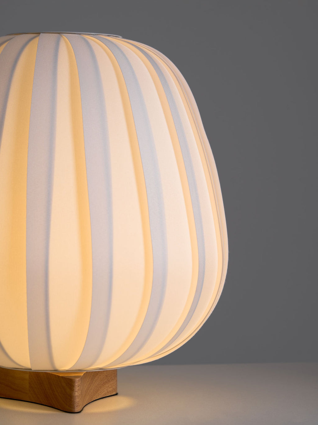 Side-on close view of a lit Clérachic table lamp, its ribbed shade glowing over a wooden base on a grey backdrop.