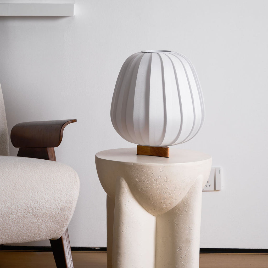 An unlit Clérachic table lamp with a white ribbed shade stands on a stone side table beside a chair with a dark wooden arm.