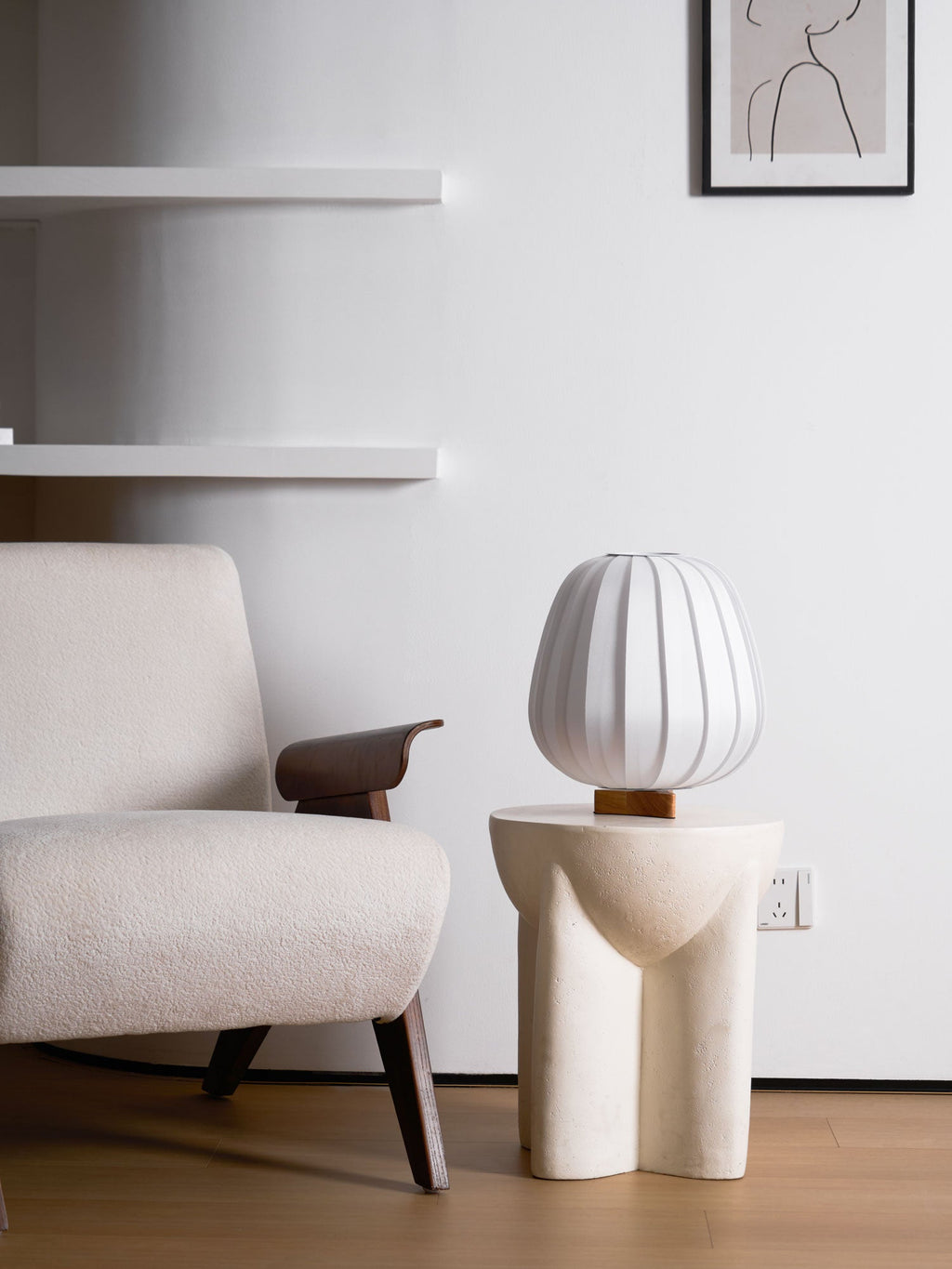 A small Clérachic table lamp stands on a stone side table beside a boucle chair, a shelf and a framed line drawing behind.
