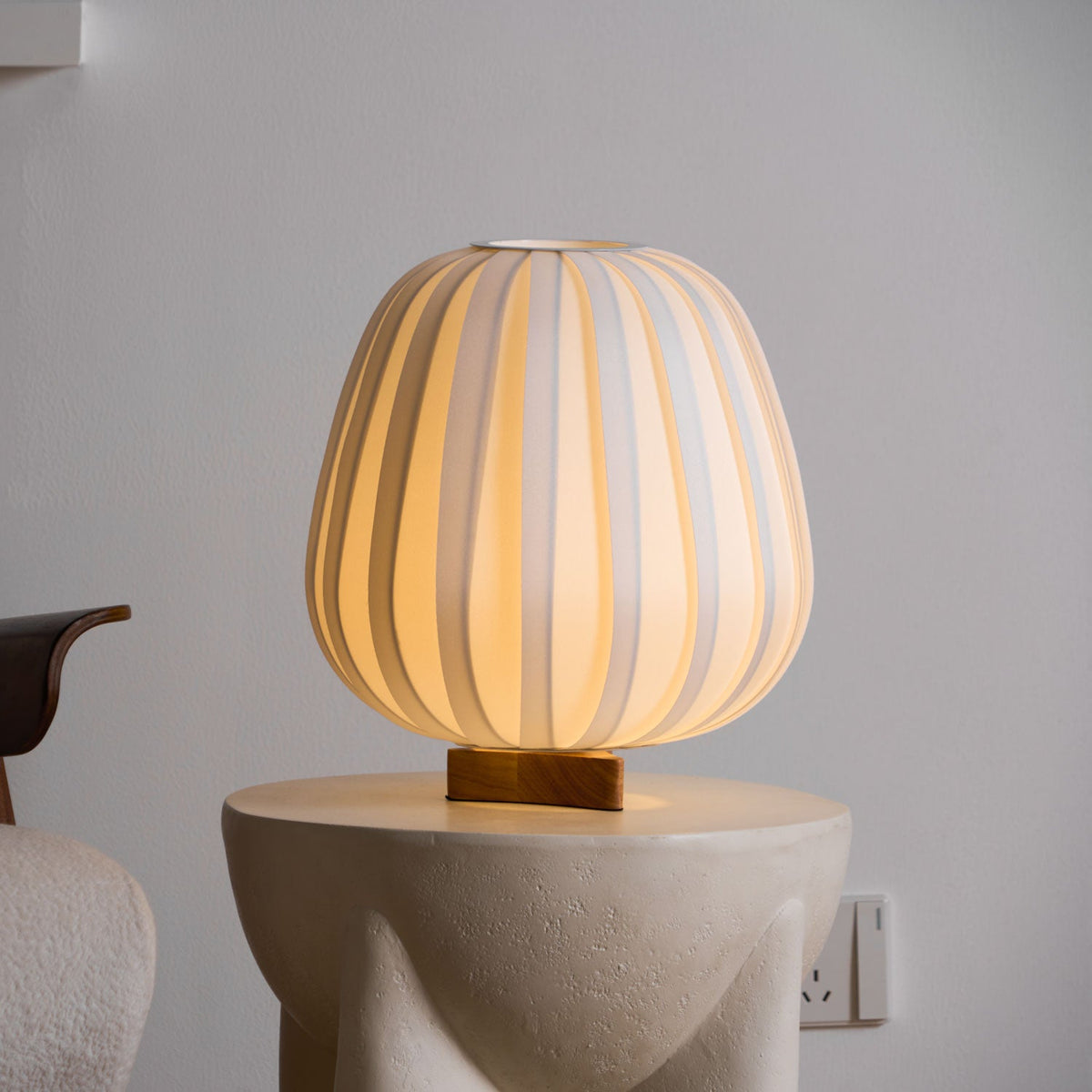 A Clérachic table lamp glows amber on a stone side table beside a chair with a dark wooden arm, against a grey wall.