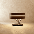 Clérachic drum table lamp with a dark wood shade, glowing band and chrome branch stem on a black base, on limewash.