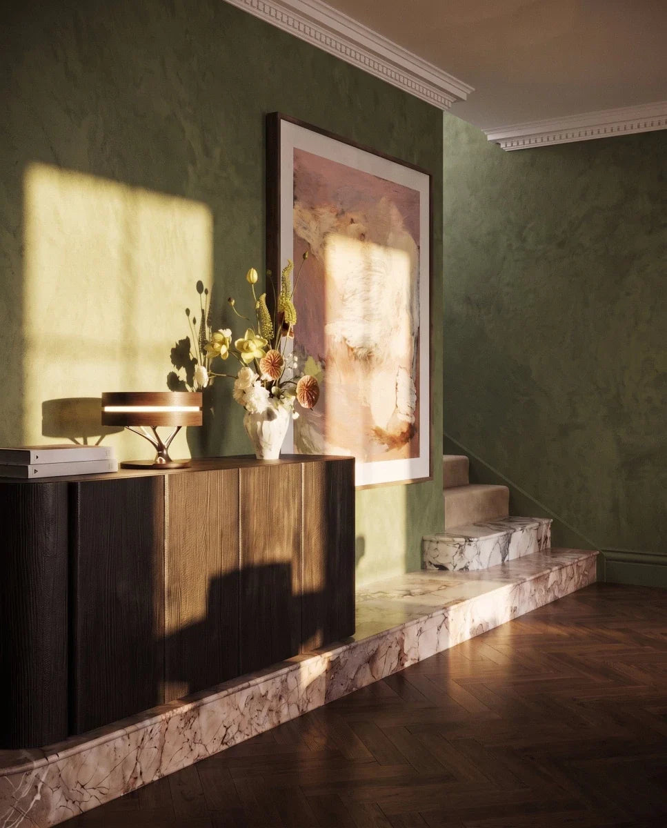 Clérachic wood-shade table lamp glowing on a dark oak sideboard beside flowers, a large print and a green plaster wall.