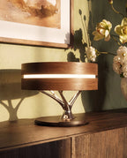 Clérachic table lamp close up on a wooden sideboard, its warm light band between two wood rings above a chrome branch stem.