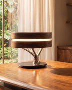 Clérachic table lamp glowing on a polished walnut desk beside sheer curtains, its chrome branch stem and black base in view.