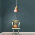 This rose-toned Clérachic cone pendant is lit against a deep teal wall above a light wooden chair.