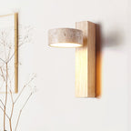 A Clérachic wall light in a pale wood finish, with a travertine head, lit softly in front of a white wall and dried twigs.