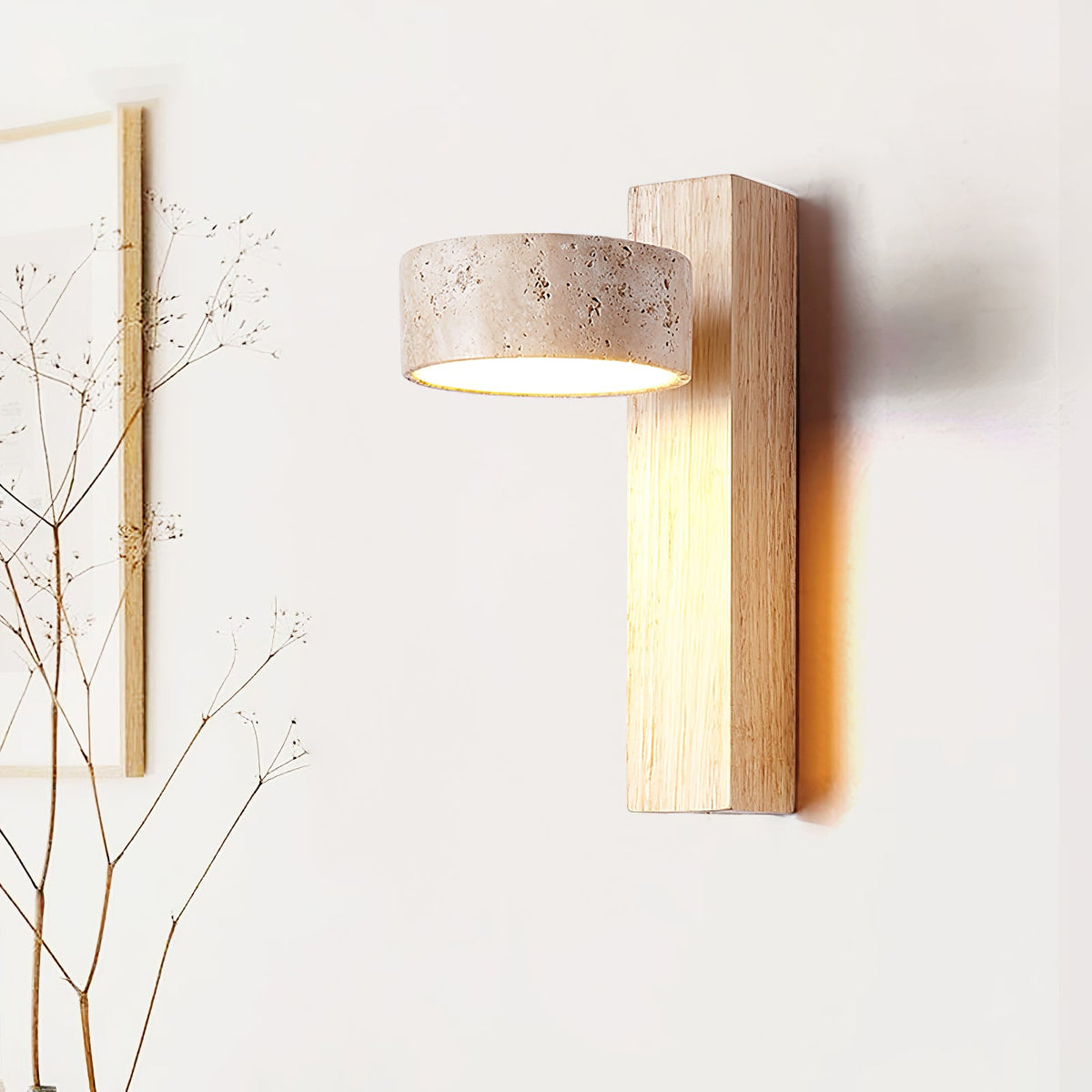 A Clérachic wall light in a pale wood finish, with a travertine head, lit softly in front of a white wall and dried twigs.
