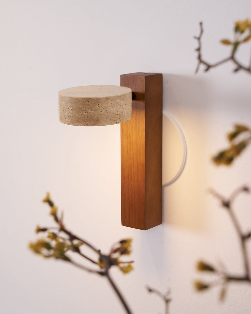 Close view of a Clérachic wall light with a walnut-coloured bar and a flat travertine head, framed by budding branches and a warm glow.