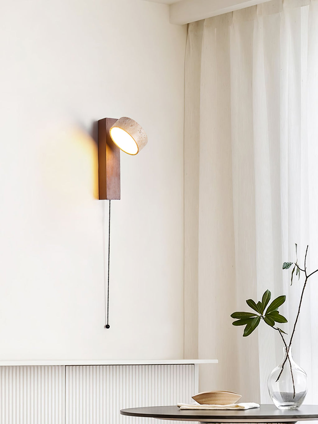 A Clérachic wall light with its head tilted sideways hangs by a curtained window, a pull cord falling past a low cabinet and a glass vase.