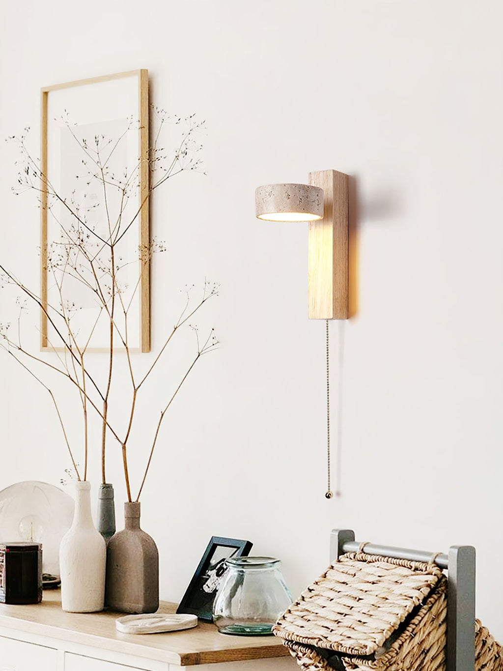 Above a sideboard with vases and a woven basket, this Clérachic wall light in pale wood has a pull cord and sits near a framed twig print.