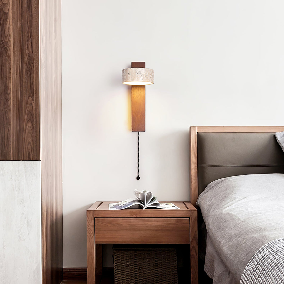 A Clérachic wall light with a walnut-coloured bar beside a bed, a pull cord hanging beside a wooden bedside table and a grey headboard.