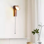 This Clérachic wall light with a walnut-coloured bar hangs by a sheer curtain, its head angled up and a pull cord falling beside a plant.