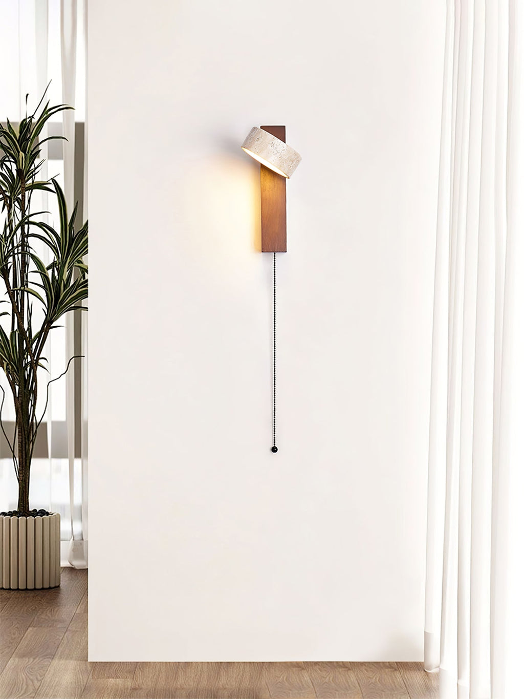 A tall white wall with a single Clérachic wall light, its head tilted and a long pull cord hanging beside a plant and a curtain.