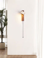 A tall white wall with a single Clérachic wall light, its head tilted and a long pull cord hanging beside a plant and a curtain.