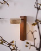 A Clérachic wall light with a walnut-coloured bar and a flat travertine head, seen through blossoming twigs against a white wall.