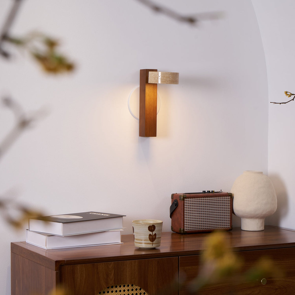 Seen through blurred twigs, this Clérachic wall light with a walnut-coloured bar sits over a sideboard holding books, a cup and a radio.