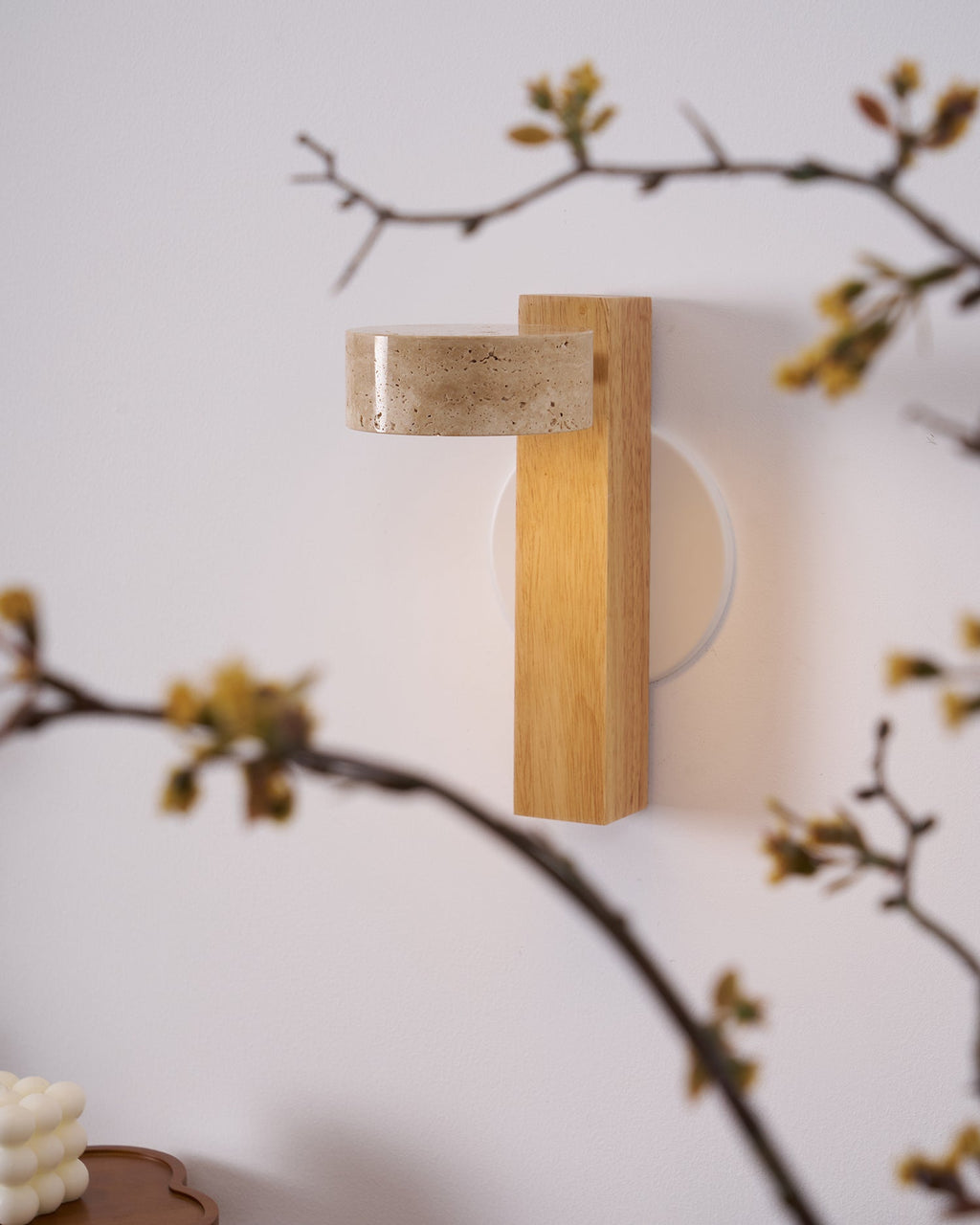 Close view of a Clérachic wall light in pale wood, its flat travertine head turned out, framed by branches with small yellow buds.