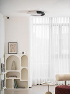 A white and black Clérachic ceiling light, shown from below, in a white room with an arched shelf and sheer curtains.